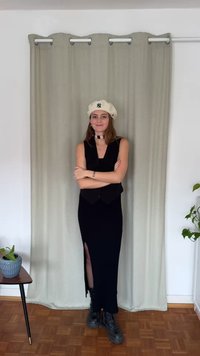 Črna obleka brez rokavov s stranskim prerezom, slojevito oblečena preko oprijete telovnika; bež baretka; črni čevlji z vezalkami; leseni tlak; zelena rastlina kot poudarek.