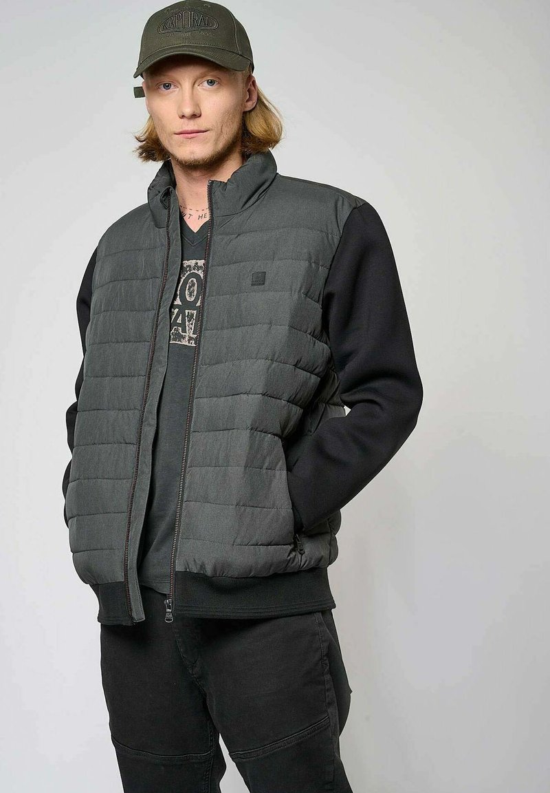 Chaqueta acolchada en gris oscuro con un cuello alto, mangas negras, bolsillos delanteros y cierre con cremallera. Lleva puesta una camiseta gráfica negra.