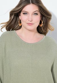 Maglione verde chiaro, texturizzato, con scollatura rotonda. Il modello indossa grandi orecchini a cerchio in tonalità oro. Comfortevole e casual, con una leggera lucentezza.