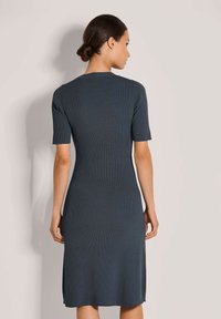 Frau mit dunklem Haar im Dutt, die ein enges, geripptes, kurzärmliges, knielanges, dunkelgraues Kleid trägt und mit dem Rücken zu einem einfarbigen hellen Hintergrund steht.