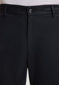 Zwarte formele broek met knoopsluiting en riemlussen, gedragen met een witte ingestopte blouse.