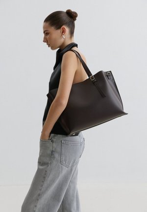 Woman with hair in a bun wearing sleeveless black top and light gray jeans, carrying large dark brown leather tote bag on shoulder.