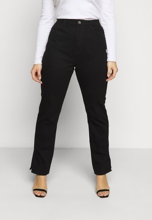 Woman wearing high-waisted black straight-leg pants with a button and zip closure, paired with black open-toe heels and a white long-sleeve top.