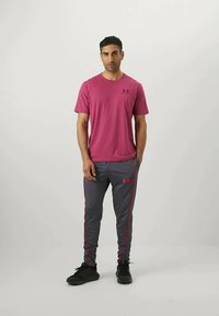 Muž stojící s rukama v kapsách, oblečený v krátkém magentovém tričku a šedých sportovních kalhotách s červenými logy Under Armour a černými botami.