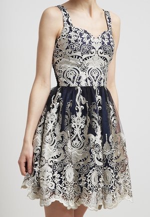 Ærmeløs mørkeblå kjole med indviklet sølvfarvet blomsterblondeoverlay og bølget kant, figursyet overdel og udvidet nederdel.