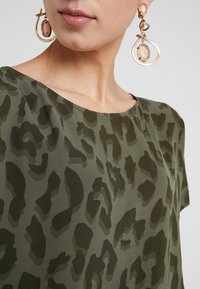 Mujer con blusa de estampado de leopardo verde y grandes pendientes colgantes dorados y de ámbar, mostrando cuello y parte inferior del rostro sobre un fondo liso.