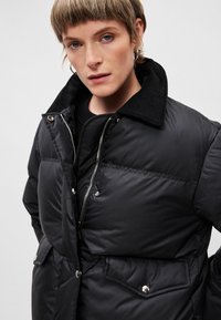 Črna napihnjena jakna z zadrgo in zapiranjem na gumb, z žametnim ovratnikom in stranskimi žepi. Gladka tekstura, izoliran dizajn.