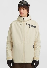 Beige waterdichte jas met een capuchon, ritszakken en zwart merkembleem. Model draagt zwarte handschoenen en een bruine beanie met een bril.
