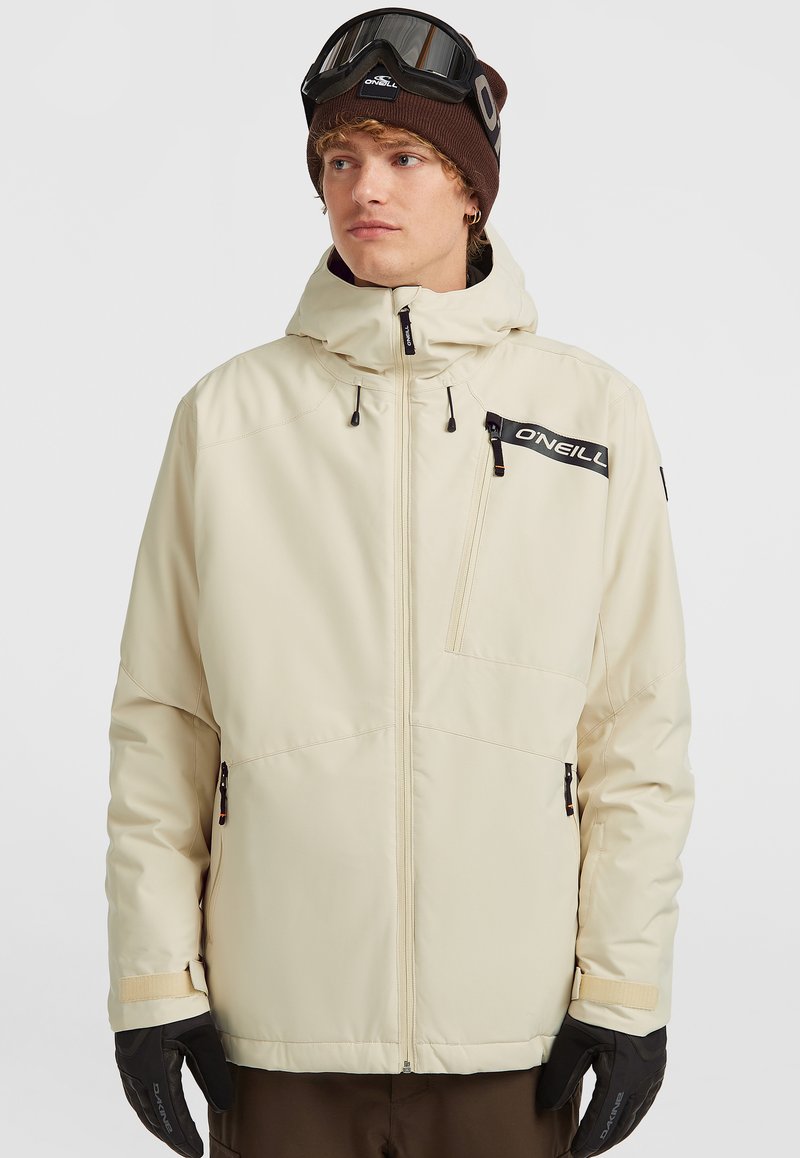 Beige waterdichte jas met een capuchon, ritszakken en zwart merkembleem. Model draagt zwarte handschoenen en een bruine beanie met een bril.