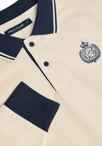 Beige Poloshirt mit marineblauen Akzenten. Verfügt über einen Kragen, eine zwei-Knopf-Leiste und eine marineblaue Brusttasche. Gesticktes Logo auf der Brust.