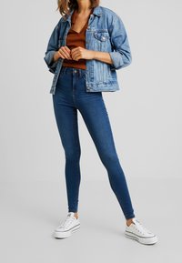 Chaqueta de mezclilla en azul claro, de mangas largas, con botones en la parte delantera, combinada con unos jeans skinny de tiro alto en azul oscuro y zapatillas blancas.