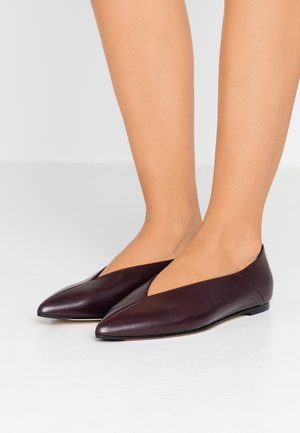 Kvinde iført mørkelilla spidse læderflade sko, der står på en hvidt gulv med bare ben synlige fra midt på læggen og ned.