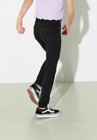 Černé skinny džíny s vysokým pasem, doplněné černobílými teniskami Vans. Džíny mají jemnou texturu a minimální detaily.