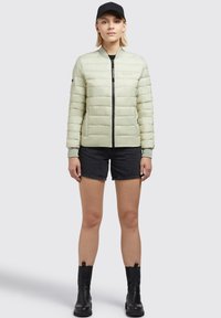 Light green puffer jacket with horizontal stitching, front zipper, ribbed cuffs, paired with black shorts and ankle boots. Black cap worn.