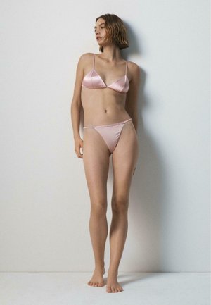 Frau steht barfuß an einer schlichten Wand, trägt einen hellrosa Satin-BH und passende Bikini-Unterwäsche und schaut leicht zur Seite.