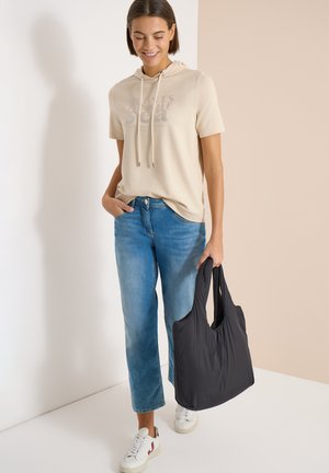 Donna che indossa una felpa con cappuccio beige a maniche corte e jeans blu, che tiene una grande borsa tote nera, in piedi contro uno sfondo neutro chiaro.