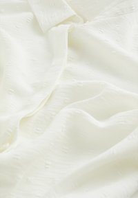 Weißer, strukturierter Stoff mit einem subtilen Muster und einem sanften Glanz, ausgestattet mit einem Knopfverschluss und einem drapierten Design. Geeignet für legere Kleidung.