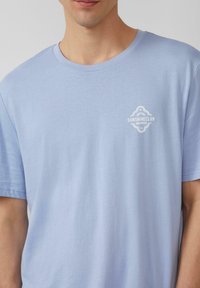 Lichtblauw katoenen T-shirt met een klein wit logo op de linkerkant van de borst, met een ronde hals en korte mouwen. Soepele textuur.