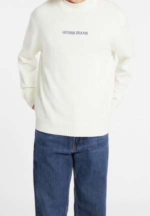Osoba nosząca biały dzianinowy sweter z napisem „GUESS JEANS” na piersi oraz niebieskie jeansy, ręce w kieszeniach swetra.