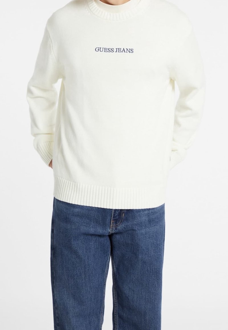 Personne portant un pull en maille blanche avec le texte « GUESS JEANS » sur la poitrine et un jean en denim bleu, mains dans les poches du pull.