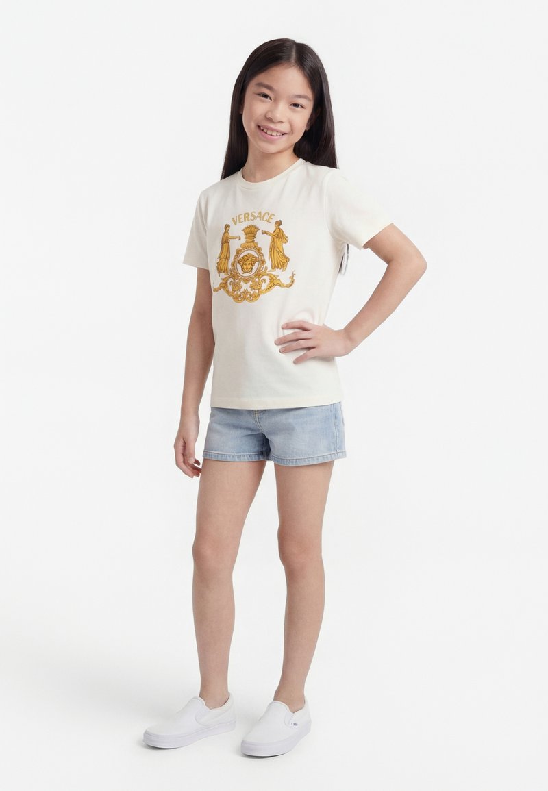 Jeune fille debout, main sur la hanche, portant un t-shirt blanc avec le logo Versace, un short en denim clair et des chaussures blanches slip-on sur fond blanc.