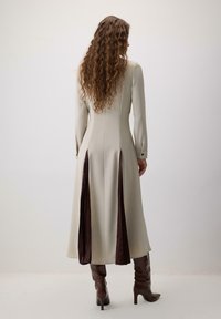 Lang, beige kjole med lange ærmer og indsnævret talje, prydet med dybe maroon sidepaneler og knaplukninger ved ærmerne, kombineret med brune hælede støvler.