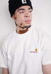 T-shirt bianco in cotone con un piccolo logo Carhartt ricamato in arancione e bordeaux sul petto, abbinato a un berretto nero.