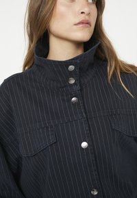 Dunkelblaues Jacket mit weißen Nadelstreifen, hohem Kragen, Druckknöpfen und zwei Brusttaschen. Der Stoff wirkt glatt und leicht strukturiert.