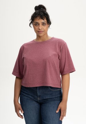 Frau mit dunklen Haaren zu einem Dutt gebunden, trägt ein lockeres kurzärmeliges mauvefarbenes Croptop und dunkelblaue Jeans, steht vor einem schlichten hellen Hintergrund.