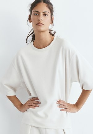 Jonge vrouw met donker haar in een losse paardenstaart, gekleed in een oversized wit T-shirt en bijpassende broek, staat met handen in de zij.