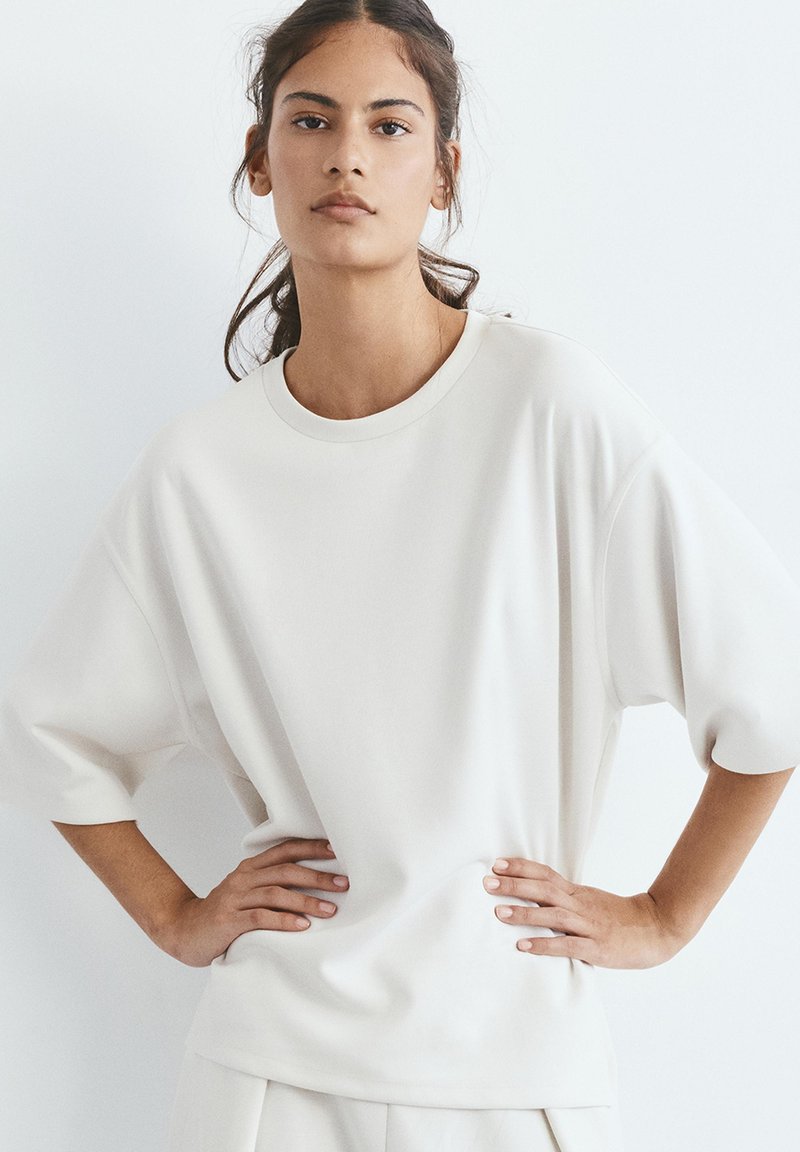 Jeune femme aux cheveux foncés en queue de cheval lâche, portant un T-shirt blanc oversize et un pantalon assorti, debout les mains sur les hanches.
