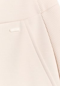 Gros plan d’un tissu beige avec un détail de manche côtelée et une petite étiquette rectangulaire cousue avec des lettres en relief.