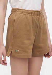 Short marron à taille élastique avec un petit logo crocodile vert sur la jambe gauche, porté avec un haut blanc.