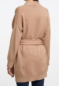 Cardigan lungo beige realizzato in un morbido tessuto con vita con cintura, polsini a coste e spacchi laterali per maggiore comfort e facilità di movimento.