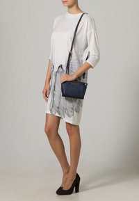 Bílé šaty s černým potiskem peří, volný střih, krátké rukávy. Tmavě modrá kabelka přes rameno s dlouhým popruhem, hladký povrch, zlaté kovové detaily.