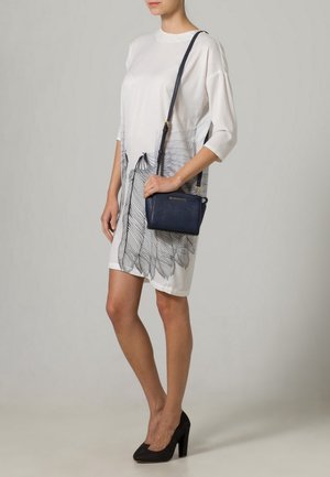 Vestito bianco con stampa di piume nere, taglio ampio, maniche corte. Borsa a tracolla blu navy con tracolla lunga, texture liscia, dettagli in oro.