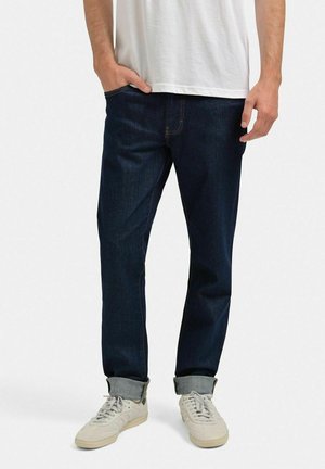 Mørkeblå jeans med lige skæring, med opbrettede kanter, kontrastsyet med gule sømme, båret med hvide sneakers og en simpel hvid t-shirt.