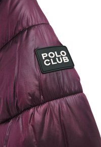 Axel på lila dunjacka med synlig sydd textur, med en rektangulär svart lapp märkt "POLO CLUB" i vita bokstäver.