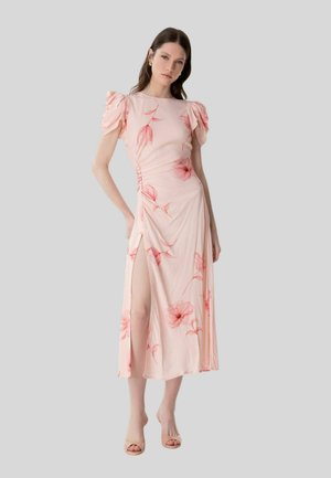 Donna che indossa un abito midi floreale rosa chiaro con maniche a sbuffo, spacco laterale e dettagli con bottoni, in piedi davanti a uno sfondo grigio semplice.