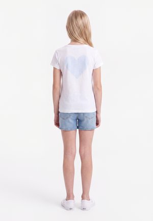 Ung pige med langt blondt hår, der står med ryggen til, iført hvid t-shirt med lyseblåt hjerte og denimshorts.
