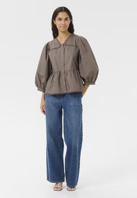 Braune Peplum-Bluse mit Puffärmeln, strukturiertem Stoff, rundem Ausschnitt und dekorativem gewelltem Saum, kombiniert mit hoch sitzenden blauen Jeans.