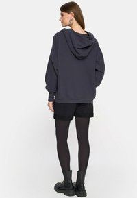 Kobieta stojąca tyłem, ubrana w ciemny, oversize'owy hoodie, czarne szorty, czarne rajstopy i czarne, masywne buty na tle gładkiego, białego tła.