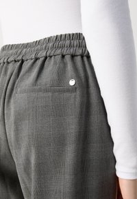 Närbild av grå rutiga byxor med elastisk midja och bakficka, som bärs med en vit långärmad topp.