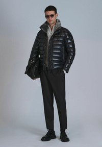 Schwarze Daunenjacke mit glänzender Oberfläche und kragen aus Fell, kombiniert mit grauem Pullover, schwarzen Hosen und schwarzen Schnürschuhen.