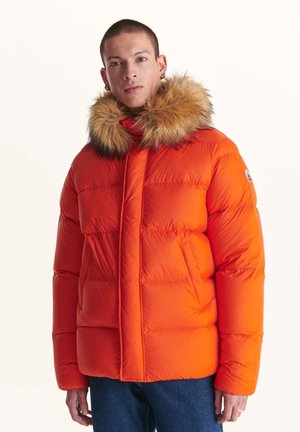 Giacca puffer arancione con cappuccio foderato in pelliccia, caratterizzata da una texture trapuntata e una chiusura frontale con zip. Due tasche laterali e patch logo sulla manica.