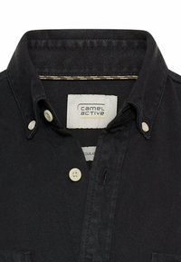 Schwarzes Jeanshemd mit Button-Down-Kragen, ausgestattet mit beigefarbenen Knöpfen, gelben Nähakzenten und einem Etikett mit der Aufschrift "camel active". Reguläre Passform.