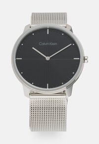 Reloj Calvin Klein con un dial circular negro, marcadores de hora plateados minimalistas, correa de malla plateada y una caja metálica delgada.