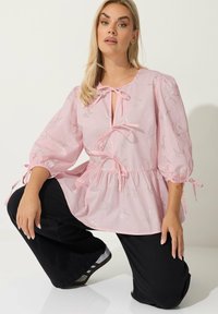 Blusa rosa con scollo a fiocco e vita arricciata, caratterizzata da motivi a fiocco argentati. Maniche lunghe a sbuffo con polsini legati. Pantaloni neri sotto.