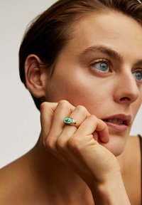 Anillo de oro con una piedra de turquesa ovalada que presenta un diseño de cruz, llevado en una mano que descansa sobre la cara, con piel clara y cabello corto.
