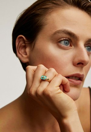 Anillo de oro con una piedra de turquesa ovalada que presenta un diseño de cruz, llevado en una mano que descansa sobre la cara, con piel clara y cabello corto.
